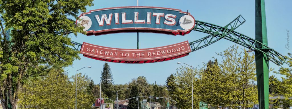 Sign reading "Willits, Gateway to the Redwoods" spans the road, with Willits urban forest in the background