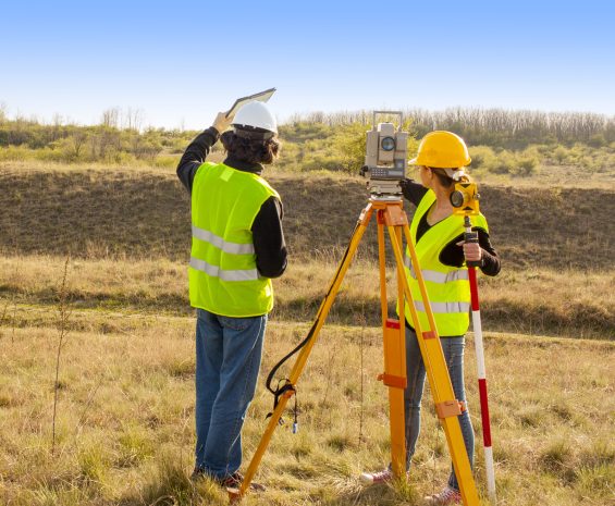 One male and one female land surveyor stand in a barren field facing into the distance. A surveying tool stands between them.