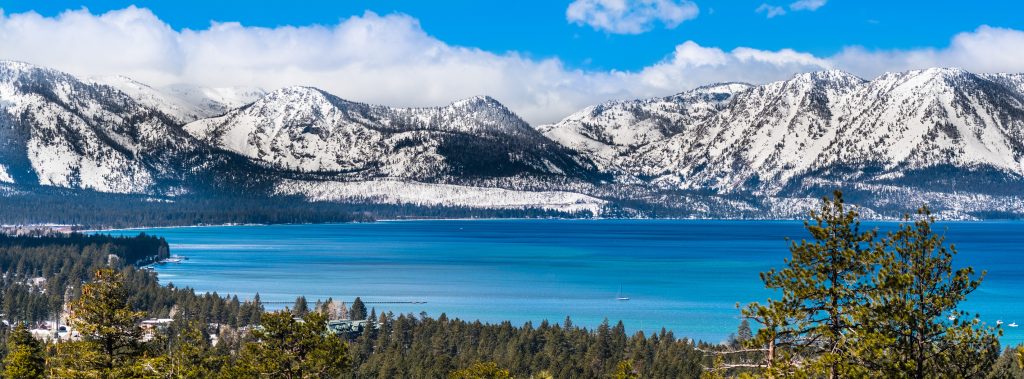 Panoramic view towards Lake Tahoe on a sunny clear day; the snow covered Sierra mountains in the background; evergreen forests in the foreground.