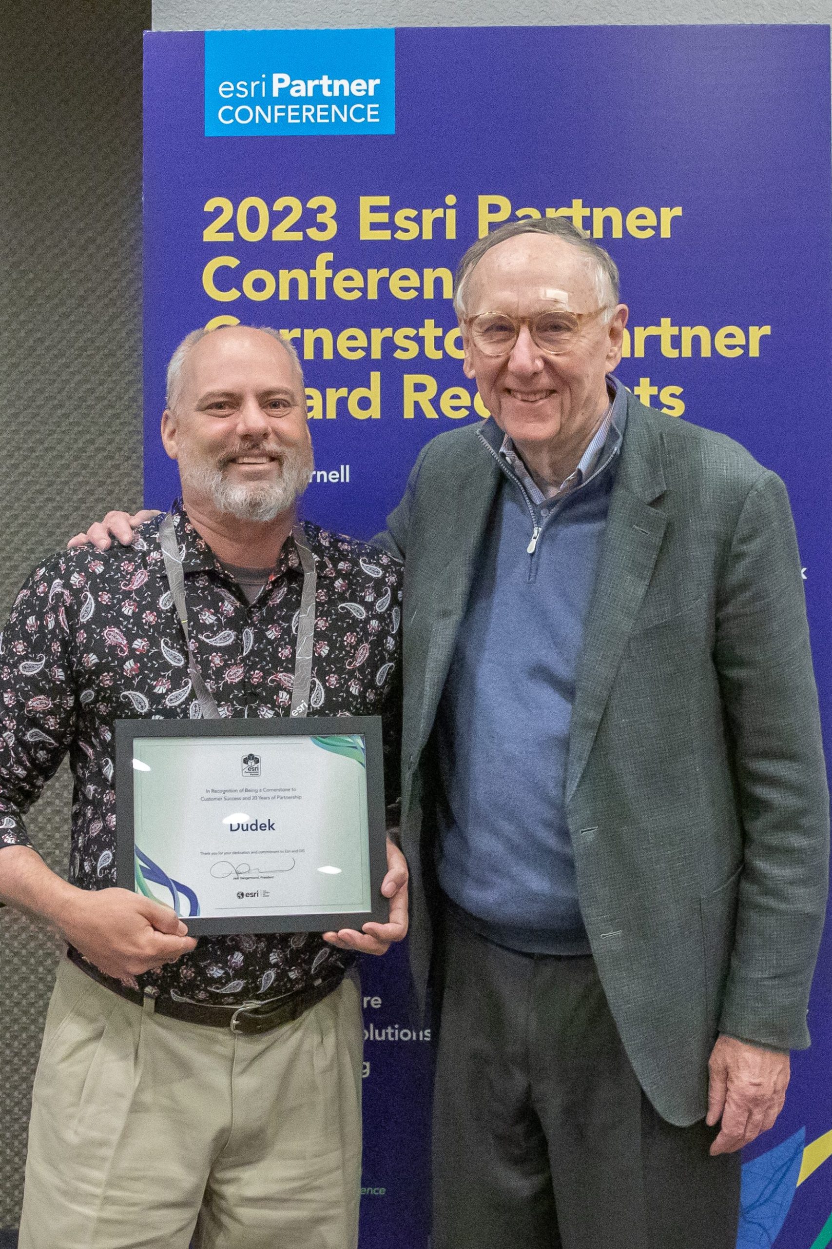 Two man stand smiling at the camera. One holds an Esri Cornerstone Partner certificate.